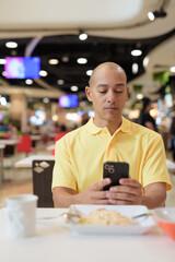 Mature bald Hispanic man using phone at indoors food court restaurant ready to eat chicken and rice