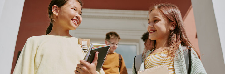 Asian girl and Caucasian girl smiling and talking while holding notebooks outdoors, standing together with two boys in background, group of children interacting after school