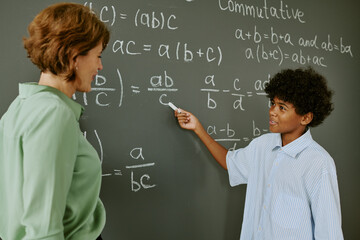Black teenage boy explaining math equation on chalkboard while middle aged Caucasian woman...