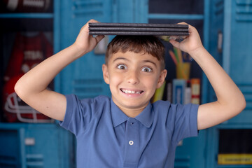 School picture of a little boy with a locker blue as a backdrop with books and pens and school supplies being silly with books on his head