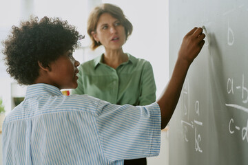 Teenage Black boy writing mathematical equations on chalkboard while middle aged Caucasian woman standing nearby observing and assisting during classroom learning activity