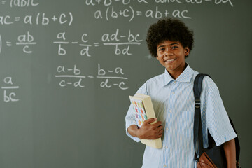 Portrait of Black teenager boy standing in front of chalkboard holding books and backpack, smiling...