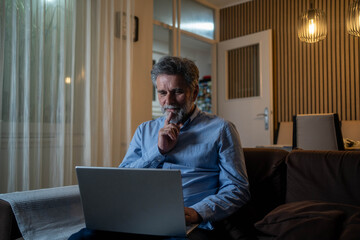 Mature man thinking while using laptop at home in living room, focused and contemplative indoors