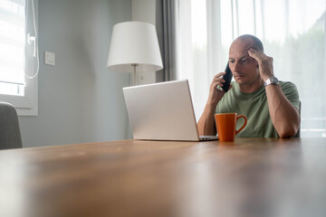 Stressed man talking on mobile phone while working on laptop at home feeling overwhelmed with...