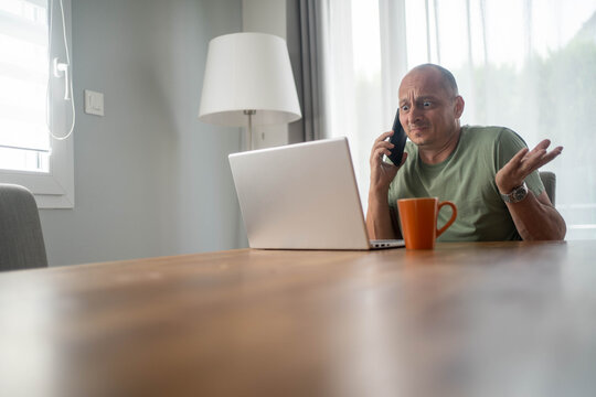 Confused man looking at laptop screen while talking on smartphone at home experiencing poor frustrating customer support