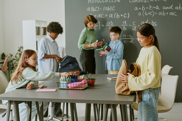 Group of multiethnic teenagers preparing for class in classroom, female teacher assisting Caucasian boy at chalkboard, Asian girl standing near table, Black boy organizing backpack