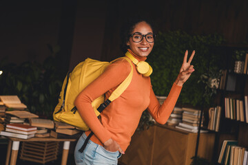 Young woman with a yellow backpack and headphones giving peace sign, surrounded by books in a cozy...