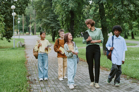 Caucasian woman walking with diverse group of children along park path, woman holding books and smiling at kids, children carrying backpacks and engaging in conversation outdoors
