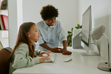 Caucasian girl sitting at desk using computer keyboard, while Black teenage boy standing beside her...