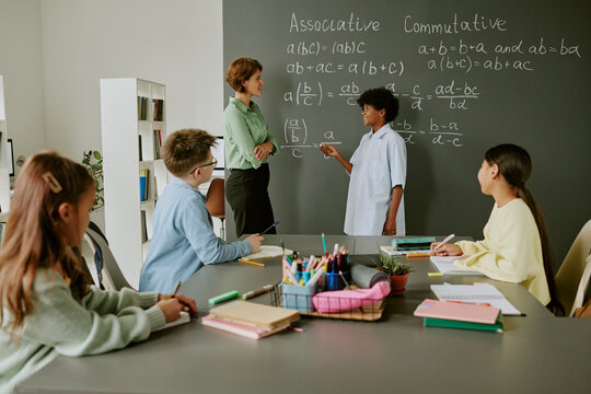 Group of diverse schoolchildren sitting at desk watching Black boy teenager standing at chalkboard solving math problem, while young Caucasian woman teacher observing and assisting in classroom