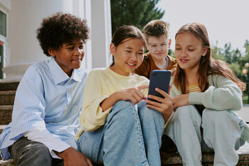 Group of multiethnic teenagers sitting outdoors using smartphone together, smiling and interacting...