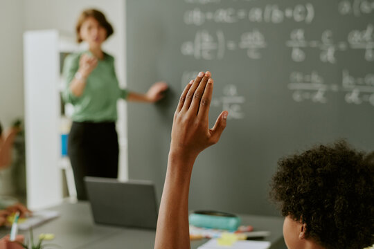 Black child raising hand in classroom while teacher standing near chalkboard with mathematical equations, engaging in lesson, visible laptop on desk, educational setting