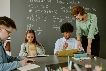 Middle aged Caucasian woman teaching diverse group of children in classroom, Black boy writing in notebook while teacher assisting, Caucasian girl and boy studying at desk during math class