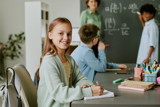 Portrait of Caucasian preteen girl smiling and writing in notebook while sitting at desk in classroom, multiethnic classmates and teacher standing near chalkboard with math equations in background