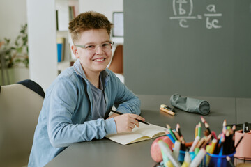 Portrait of Caucasian boy smiling while sitting at desk and writing in notebook, looking into camera, surrounded by school supplies, chalkboard with math equations in background, classroom setting