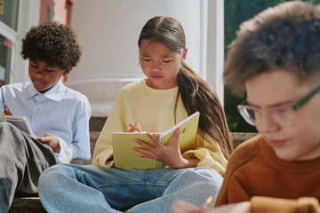Multiethnic group of preteen children sitting outdoors writing in notebooks, Asian girl concentrating on homework, Caucasian boy and Black boy studying together on steps