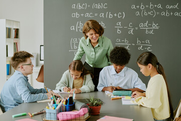Middle aged Caucasian woman teaching diverse group of children sitting at table and writing in classroom with chalkboard covered in math equations, students focused on assignments