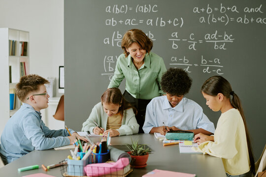 Middle aged Caucasian woman teaching diverse group of children sitting at table and writing in classroom with chalkboard covered in math equations, students focused on assignments