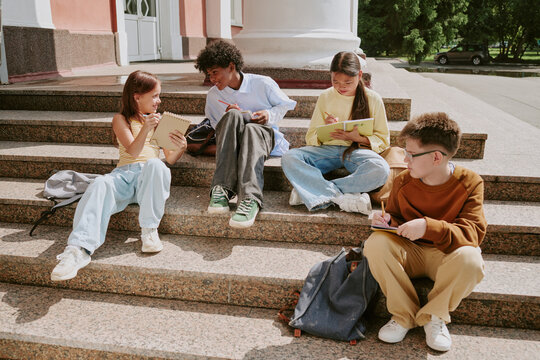 Group of multiethnic teenagers sitting on outdoor steps writing in notebooks and talking, showing diverse group of boys and girls engaged in collaborative learning activity together