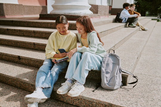 Two teenage girls, one Asian and one Caucasian, sitting on outdoor steps sharing smartphone and books, smiling and interacting, with group of diverse teenagers talking in background