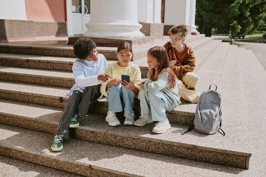 Group of multiethnic children sitting on outdoor steps using smartphone together, two girls and two boys interacting and smiling, backpack placed nearby, casual setting