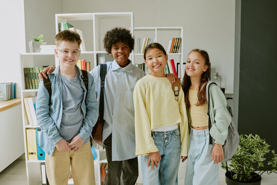 Group of diverse teenagers standing together smiling at camera, multiethnic boys and girls with backpacks posing in classroom setting, bookshelves in background