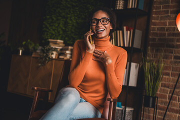 Young woman in orange turtleneck sweater enjoys a pleasant phone call while sitting indoors.