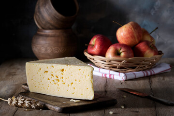 Piece of cheese on cutting board and apples in wicker plate on rustic table