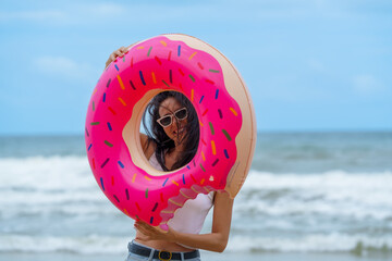 Woman enjoys sunny day at beach with friends, playfully holding large donut shaped float while sea...