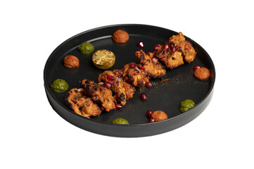 Classic chicken tikka marinated in yoghurt and spices, skewered and cooked in a clay oven, served with green and red chutney, garnished with anardana on a black plate.