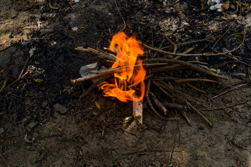 A cozy campfire glows in a rustic outdoor scene, with orange and yellow flames dancing among dry sticks and twigs arranged in a teepee. Surrounded by scattered debris and a plastic bag, this impromptu