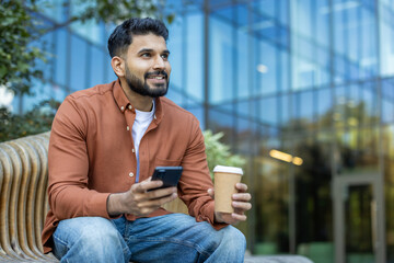 Smiling man with coffee sitting outdoors using smartphone in an urban area