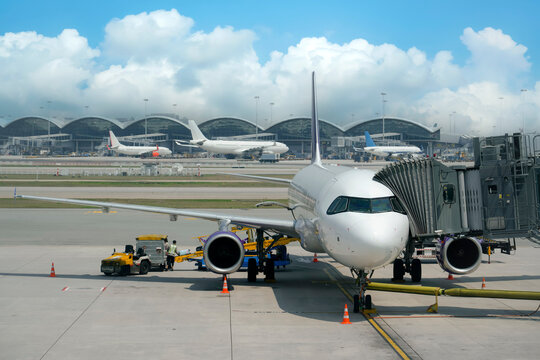 Passenger airplane is parked on the tarmac at Hong Kong International Airport. A boarding bridge connects to the plane. Airport staff work nearby on the ground with ground vehicles - Powered by Adobe