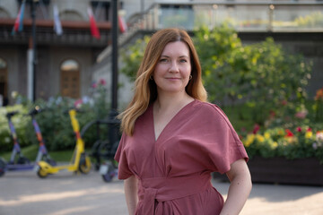 Fototapeta premium Confident middle-aged woman in a rose jumpsuit stands outdoors with colorful flowers and e-scooters in the background
