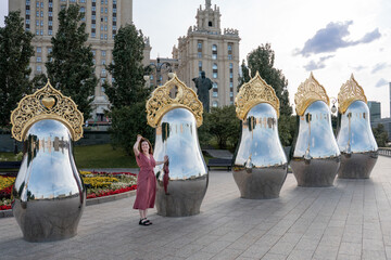 Woman in a rose dress stands beside giant reflective matryoshka sculptures with ornate golden crowns at Taras Shevchenko Embankment in Moscow