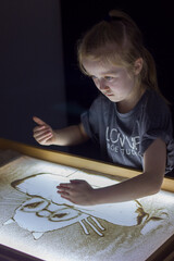 Beautiful little girl draws with sand on a light square, art therapy, sand animation, creative leisure for children, selective focus