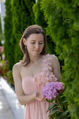 Fototapeta premium Young woman in an elegant light pink dress with floral details gently holding a blooming purple hydrangea, standing beside lush green bushes in a summer garden
