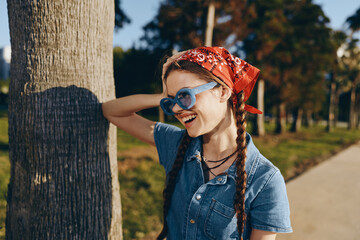 smiling woman with braids wearing sunglasses and red bandana standing by tree in sunny park, casual...