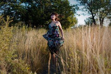 Woman strolls through tall grass in a sunlit meadow, wearing sleeveless floral dress that...