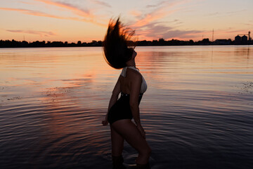 Fototapeta premium Woman in black bikini spins in shallow water at sunset, hair flowing freely as she faces away from camera. Calm lake reflects warm sky, with silhouetted trees and industrial structures in background