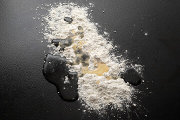 A pile of scattered flour and vegetable oil on a black background. Top view. Making dough from...