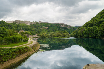Obraz premium Viewpoint across the Doubs river with Citadelle de Besançon above lush hills, ideal for travel editorials, French heritage, nature, and city tourism themes.