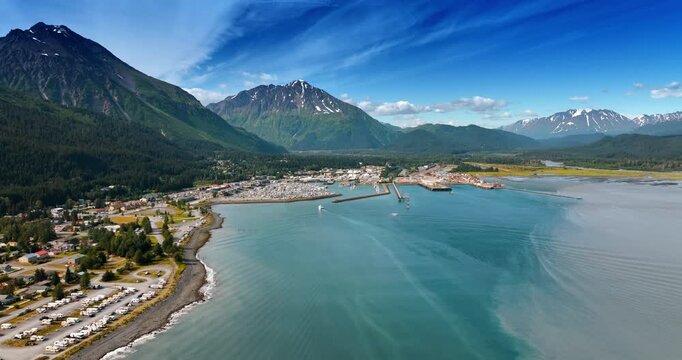 Flight over the blue waterscape of the lake near the town located on the beautiful waterfront. Evergreen forests and stunning mountains surround the urban area. Alaska, aerial view.