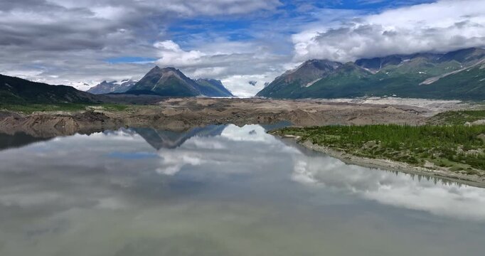 Stunning overcast sky reflects in the smooth surface of the lake. Drone rising slowly over the waterscape revealing the view on the gorgeous mountains. Alaska wilderness.