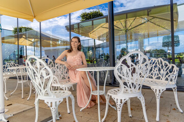 Beautiful young woman in a pink dress with floral details sitting at a white wrought iron table in...