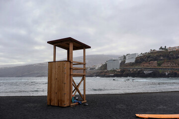 A picturesque wooden lifeguard tower stands on a black sand beach beside a calm ocean, creating a...