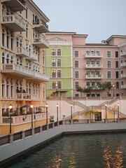 A vibrant canal flows past colorful European-style buildings with ornate balconies and lit streetlights at dusk.