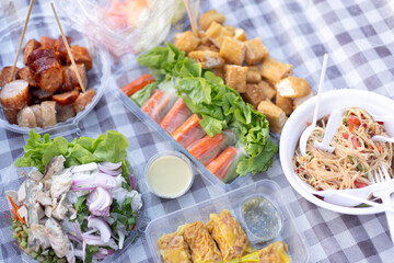 A casual outdoor picnic setup with a variety of Thai dishes , Mackerel Spicy Salad , Pork Dumpling , Spicy Rice Vermicelli Salad , fresh vegetables, Grilled Sausage , and spring rolls.