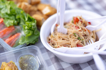 A casual outdoor picnic setup with a variety of Thai dishes , Mackerel Spicy Salad , Pork Dumpling , Spicy Rice Vermicelli Salad , fresh vegetables, Grilled Sausage , and spring rolls.