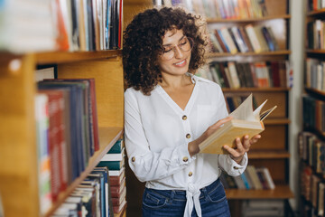 Young student reading a book in the library surrounded by bookshelves